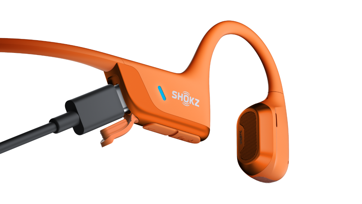 An orange bone-conduction sport earpiece with a black charging connector attached, shown on a clean white background. The brand name 'SHOKZ' is printed on the side.