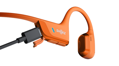 An orange bone-conduction sport earpiece with a black charging connector attached, shown on a clean white background. The brand name 'SHOKZ' is printed on the side.